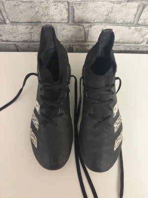 Svarta Adidas predator fotbollskot (storlek 36) - Säljer ett par svarta Adidas fotbollsskor med vita ränder och coolt dropp-mönster på sidorna. Skorna har snörning, platt sula med dobbar och är tillverkade i syntetmaterial med stickad ovandel för extra komfort. Perfekt för dig som vill ha stil på planen.