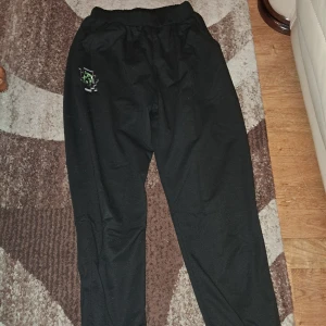 Svarta mjukisbyxor med Minecraft Creeper - Svarta mjukisbyxor med elastisk midja och muddar vid benslut. På ena benet finns ett tryck med en pixelgrafik av en Creeper från Minecraft som ser ut att spränga sig ut ur tyget. Sköna och avslappnade, perfekta för gaming eller chill. Materialet känns som bomull.