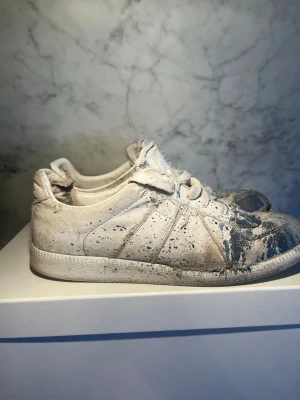 Vita Maison marigela gats med blåt färgstänk - Unika vita sneakers med coola färgstänk i blått. Skorna har en låg profil, rund tå och snörning. Materialet är canvas med tydliga slitningar och fransiga detaljer som ger en rå och edgy vibe. Perfekt för dig som gillar streetstyle och vill sticka ut. På en av bilderna ser dem rätt slitna ut men det är bara att Lima så är det bra igen hör av vid frågor om och bilder om skorna!!