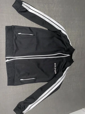 Palm Angels svart tracksuit med vita ränder - Svart tracksuit från Palm Angels med vita ränder längs ärmar och ben. Setet består av en zip-jacka med två fickor och matchande byxor med elastisk midja. Palm Angels-logga broderad på både jacka och byxa. Tillverkad i mjuk polyester för en sportig vibe.