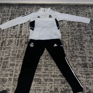 Vit Adidas Real Madrid tracksuit - Snygg vit långärmad träningströja från Adidas med Real Madrid-logga på bröstet. Tröjan har svart detalj på sidorna, half zip och klassisk Adidas-logga. Perfekt för fotbollsträning eller chill. Materialet är polyester och passformen är normal.