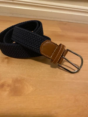 Mörkblå flätat bälte med skinn - Snyggt mörkblått flätat bälte med bruna skinn-detaljer och silvrigt spänne. Bältet har en stilren och modern look med tydlig struktur i materialet. Perfekt för att ge outfiten en extra touch. Passar till jeans eller chinos.