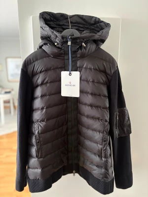 Svart Moncler padded zip-up cardigan  - Snygg svart cardigan från Moncler med stickade ärmar och quiltad framsida. Jackan har en stor huva, dragkedja framtill och Moncler-logga på ärmen.  Aldrig använd, säljes pga fel storlek. 