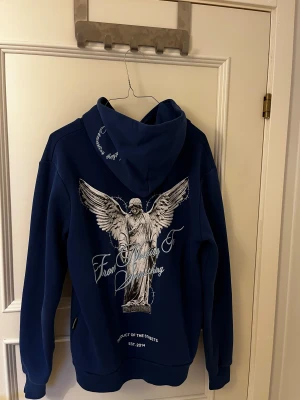 Blå hoodie från Hoodrich med tryck - Säljer en blå hoodie från Hoodrich i storlek S. Tröjan har en stor vit ängel- och textprint på ryggen samt mindre tryck framtill. Klassisk huva och känguruficka. Perfekt för dig som gillar streetwear och snygga detaljer.