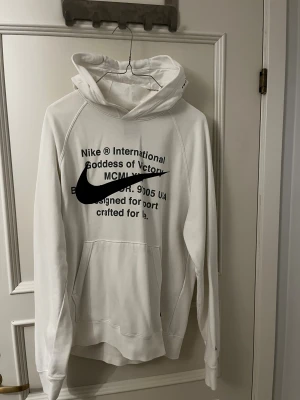 Vit Nike hoodie - Vit hoodie från Nike i storlek M med svart tryck och stor Swoosh-logga på framsidan. Tröjan har huva, känguruficka och långa ärmar. Materialet är mjuk bomull och polyester, perfekt för chill eller sport. Snyggt minimalistiskt utseende med textdetaljer framtill.