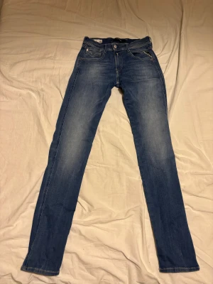 Replay Anbass Hyperflex jeans blå 28/32 - Slim fit jeans från Replay, modell Anbass Hyperflex Recycled 360°. Snyggt blå tvätt med klassiska fem fickor och vita Replay-loggan på bakfickan. Stretchigt material för extra komfort och smidig passform. Perfekt för dig som gillar stilrena och bekväma jeans.