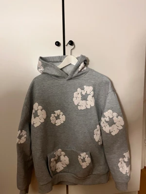 Grå hoodie med vita blommor Denim Tears - Grå hoodie från Denim Tears med vita blommor tryckta över hela tröjan. Tröjan har huva, magficka och ribbade muddar. Mjuk insida i fleece som gör den extra skön. Perfekt för dig som gillar streetwear och vill sticka ut med ett unikt mönster.