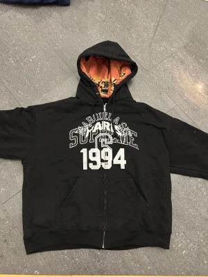 Supreme x Margiela svart hoodie 1994 - Svart hoodie från Supreme x Margiela med stor vit print 'SUPREME 1994' på bröstet. Insidan av huvan har ett fett döskallemönster i orange, gul och svart. Tröjan har snörning i huvan, dragkedja framtill och känguruficka. Perfekt för dig som gillar streetwear med edge.