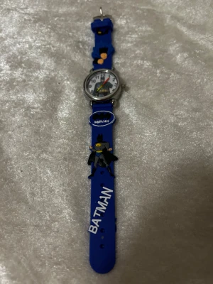  Batman barn klocka med motiv - Säljer en cool blå Batman-klocka med motiv av Batman på både urtavlan och armbandet. Klockan har ett silverfärgat runt ur och färgglada visare. Armbandet är i syntetmaterial med tydliga Batman-detaljer och text. Perfekt för dig som gillar superhjältar! 3 år + 