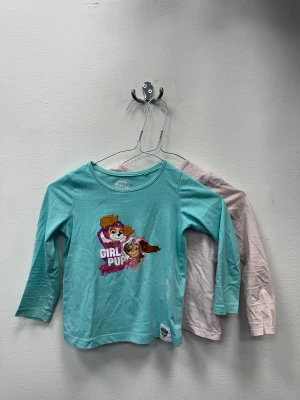 2-pack långärmade tröjor Paw Patrol - Två långärmade tröjor i mjuk bomull. Ena tröjan är turkos med tryck av Skye och Everest från Paw Patrol och texten 'Girl Pup Power' i rosa och vitt. Den andra tröjan är enfärgad ljusrosa. Båda har rund hals och normal passform.