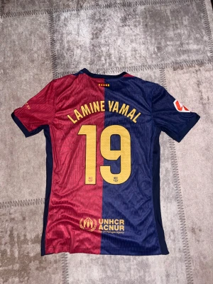 FC Barcelona Lamine Yamal matchtröja - Säljer en officiell FC Barcelona matchtröja från Nike med Lamine Yamal och nummer 19 på ryggen. Tröjan har halva sidan vinröd och halva marinblå, med gul text och detaljer. Kortärmad, med FCB-emblem och Dri-FIT ADV material för bästa komfort.
