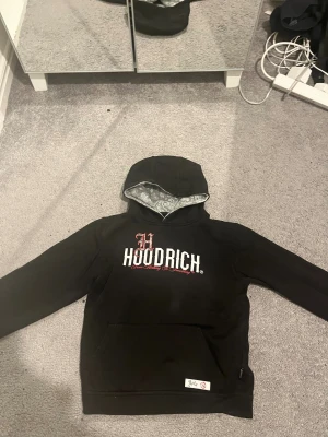 Svart hoodie från Hoodrich med paisley - Svart hoodie från Hoodrich med stor logga på bröstet och paisleymönstrad insida i huvan. Snygga röda och vita detaljer i trycket, samt patch vid fickan. Klassisk känguruficka och långärmad modell, perfekt för streetwear-stilen.