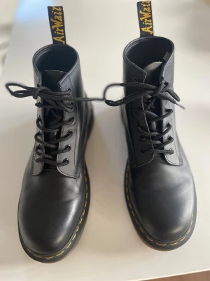 Fynda Svarta Dr. Martens orginal boots i skinn - Säljer ett par klassiska svarta Dr. Martens boots med rund tå och snörning. Skorna har gul söm runt sulan och tydlig AirWair-flik bak. Tillverkade i slätt skinn med robust platt sula i gummi. Nästintill oanvända och ett perfekt fynd för vintern. Storlek 43