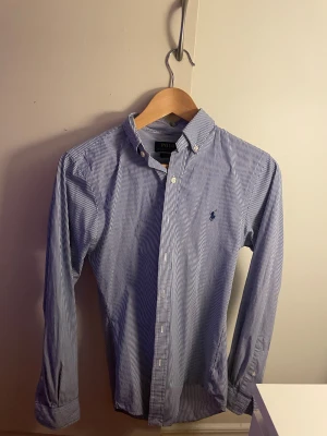 Blårandig skjorta från Polo Ralph Lauren - Snygg blårandig skjorta från Polo Ralph Lauren med klassisk button-down krage och broderad logga på bröstet. Skjortan har långa ärmar, vita knappar och är gjord i mjuk bomull. Perfekt för dig som gillar stilrena och tidlösa plagg.