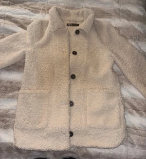 Beige teddyjacka från Zara XS - Säljer en beige teddyjacka från Zara i storlek XS. Jackan har stora framfickor, krage och svarta knappar framtill. Den är fluffig och har en loose passform, perfekt för dig som gillar mysiga och trendiga jackor.