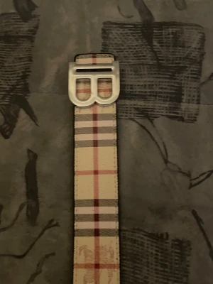 Beige och rutig Burberry bälte - Snyggt bälte från Burberry med klassiskt rutigt mönster i beige, rött och grått. Bältet har en stilren silverfärgad metallspänne med Burberrys ikoniska B-form. Perfekt accessoar för att lyfta din outfit med lite lyxig känsla.