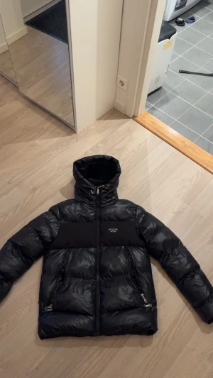 Svart pufferjacka från Alessandro zavetti Milano - Svart pufferjacka från Alessandro zavetti Milano med glansig finish och breda paneler. Jackan har huva med snörning, två sidofickor med dragkedja och en snygg logga på bröstet. Perfekt för kalla dagar och har en modern, urban vibe. Väldigt bra skick, en liten sömn på men den syns knappast eftersom jackan är svart!