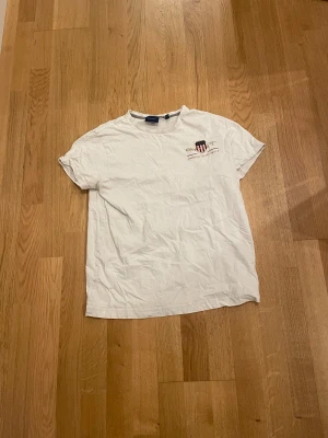 Vit t-shirt från GANT med tryck - Vit t-shirt från GANT i storlek S med klassiskt tryck och logga på bröstet. Rund hals och korta ärmar, tillverkad i mjuk bomull. Enkel och clean design med liten emblem-detalj framtill. Perfekt för en chill och stilren look.