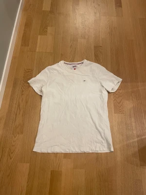 Vit t-shirt från Tommy Hilfiger - Vit t-shirt från Tommy Hilfiger med klassisk rund hals och liten broderad logga på bröstet. T-shirten är kortärmad och har en clean, enkel design som funkar till allt. Tillverkad i mjuk bomull för skön känsla hela dagen.