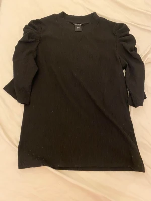 Svart T-shirt med puffärm från Lindex - Svart kort klänning från Lindex med snygga puffärmar och struktur i tyget. Modellen har rund halsringning och trekvartsärm. Perfekt för dig som gillar stilrena plagg med en twist. Materialet känns mjukt och har en lätt ribbad känsla.