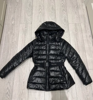 Svart vinterjacka från Calvin Klein - Snygg svart vinterjacka från Calvin Klein med huva och bälte i midjan med logga. Jackan har quiltad design, dragkedja framtill och två fickor med dragkedja. Perfekt för kalla dagar och ger en fin form med sin lite längre passform och bälte i midjan 