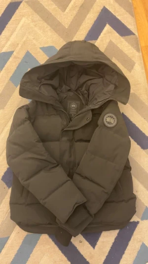  Canada Goose jacka - Svart pufferjacka från Canada Goose med stor huva och ikonisk patch på ärmen. Jackan har dragkedja och knappar framtill, samt quiltad design som ger extra värme. Perfekt för kalla dagar och har en clean, stilren look.