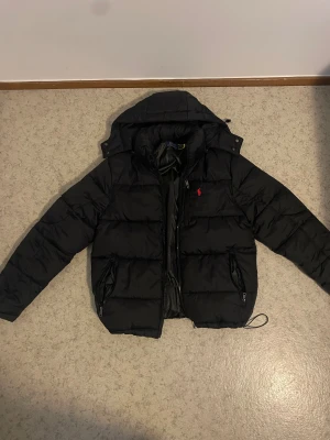 Svart pufferjacka från Polo Ralph Lauren - Säljer en snygg och klassisk svart dunjacka från Polo Ralph Lauren i mycket bra skick. Jackan är välvadderad och håller värmen riktigt bra under kalla dagar. Den har en avtagbar/justerbar huva, två rymliga ytterfickor med dragkedja samt en praktisk innerficka.