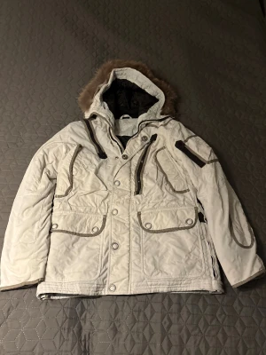 Vit dunjacka med päls från First Slope - Säljer en vit dunjacka från First Slope med beige detaljer och päls på huvan. Använd fåtal gånger!!❤️Jackan har flera fickor med dragkedja och knappar, broderad logga på ärmen och insidan, samt quiltad design. Perfekt för kalla dagar med sin tjocka och varma känsla.