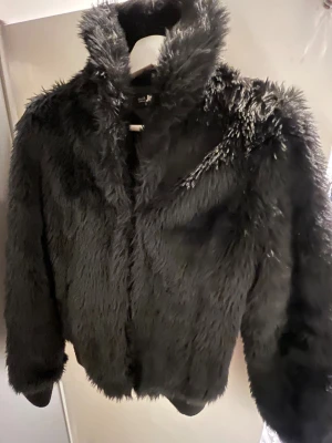 Svart pälsjacka med fluffig look - Säljer en svart pälsjacka med riktigt fluffig och mjuk känsla. Jackan har hög krage och långa ärmar, inga fickor. Knäpps med husker. Inköpt på JC.