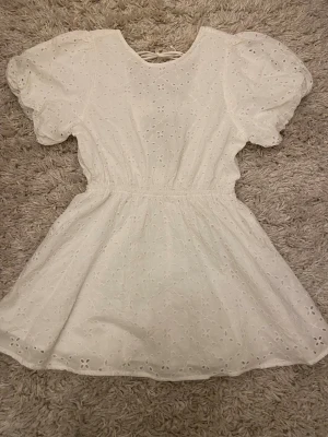 Vit kort klänning från Gina Tricot - Supersöt vit kort klänning från Gina Tricot med broderade blommor, puffärmar och två knytband i ryggen. Klänningen har markerad midja och är tillverkad i bomull, perfekt för sommarens alla tillfällen. Snygg och luftig med fina detaljer!