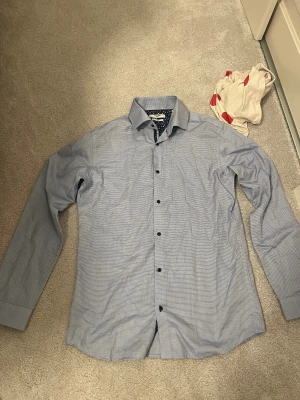 Blå slim fit skjorta Jack & Jones - Slim fit skjorta från Jack & Jones i ljusblått med diskret rutigt mönster. Insidan av kragen har ett snyggt blommigt tyg i mörkblått och vitt. Skjortan har svarta knappar och klassisk krage, perfekt för dig som gillar stilrena detaljer. Använd 1 gång i cirka 45min 