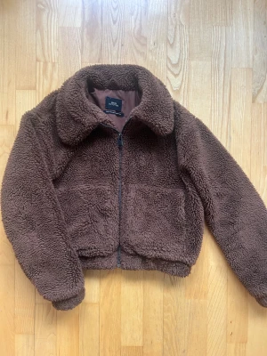 Brun teddyjacka från Urban Outfitters - Mysig brun teddyjacka från Urban Outfitters med fluffigt material och croppad passform. Jackan har en klassisk krage, två stora framfickor och dragkedja framtill. Perfekt för att hålla värmen under kyliga dagar. Nypris 800kr
