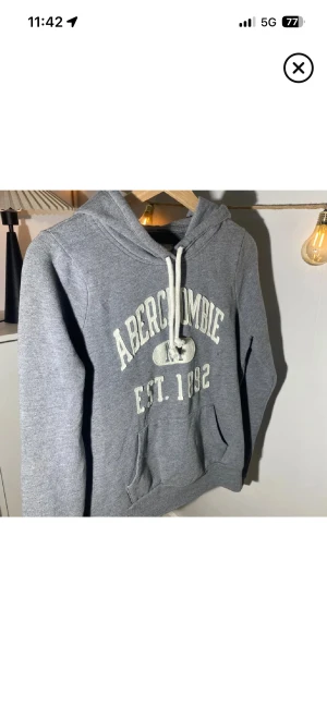 Grå hoodie Abercrombie NY - Snygg grå hoodie från Abercrombie & Fitch med vit text 'Abercrombie NY EST. 1892' på bröstet. Tröjan har huva med snörning, magficka och långa ärmar. Perfekt för chill dagar och streetstyle. Mjuk och skön bomullsblandning.  VÄLDIGT GRISH 
