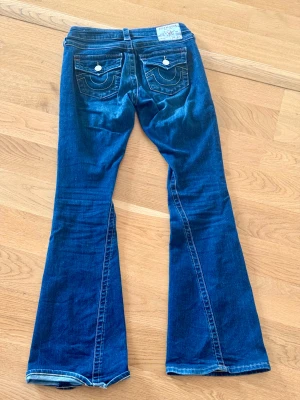 True Religion jeans  - Snygga och klassiska True Religion-jeans i modellen Joey Low Rise Flare, storlek 25. Dessa jeans har den ikoniska True Religion-designen med snygga detaljer på bakfickorna och utsvängda ben som ger en riktigt flatterande passform.                            • Modell: Joey – Low Rise Flare • Storlek: 25 • Färg: Mörk denim • Äkta True Religion (se lapparna på bilderna) • Fint använt skick med naturliga spår av användning