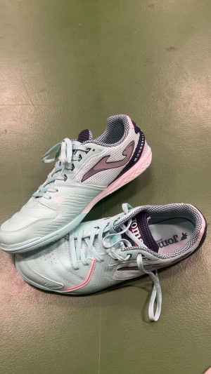 Mintgröna fotbollsskor från Joma - Snygga mintgröna fotbollsskor från Joma med ljusblå snörning och detaljer i rosa och mörkblått. Skorna har rund tå, platt sula och ventilerande mesh vid hälen. Perfekt för dig som vill sticka ut på planen med stil.