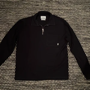 Marinblå half zip tröja från Woodbird - Marinblå långärmad tröja från Woodbird med krage och half zip-dragkedja framtill. Tröjan har en bröstficka med liten etikett och ribbade muddar vid ärmslut. Tillverkad i ekologisk bomull för en skön och clean look.