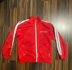 Röd trackjacka från Palm Angels - Palm Angels trackjacka i rött med vita och svarta ränder längs ärmarna. Jackan har hög krage, hel dragkedja framtill och två fickor med dragkedja. Loggan är tryckt på bröstet. Perfekt för en sportig och streetwear-inspirerad look.