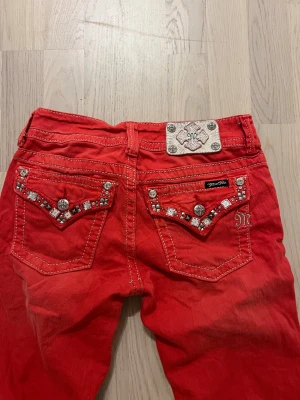 Röda Miss Me skinny jeans med nitar - Snygga röda skinny jeans från Miss Me med vita kontrastsömmar och dekorerade fickor med nitar och strass. Byxorna har låg midja och klassisk femficksdesign. Märkeslogga på bakfickan och metallmärke i midjan. Perfekta för dig som vill sticka ut.