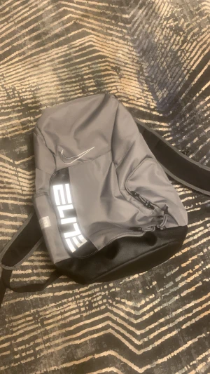 Grå Nike Elite ryggsäck - Snygg grå Nike Elite ryggsäck med svart botten och vita detaljer. Väskan har flera fack med dragkedja, vadderade axelremmar med transparenta paneler och stor ELITE-logga. Perfekt för att bära allt du behöver till skolan eller träningen.