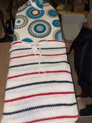 Randig stickad tröja med snörning - Unik stickad tröja i vitt med röda och mörkblå ränder, handgjord av Anna Jönsson. Tröjan har långärmade ärmar och snörning vid halsen för en cool detalj. Perfekt för dig som gillar retrovibbar och vill ha något ingen annan har.priset kan diskuteras