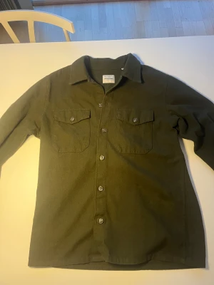 Olivgrön overshirt från Jack & Jones - Olivgrön overshirt från Jack & Jones med klassisk krage och två bröstfickor med knappar. Skjortan har långa ärmar och stängs med knappar framtill. Materialet är mjukt och känns som bomull, perfekt för lager-på-lager. Snygg och enkel att styla till jeans eller chinos.