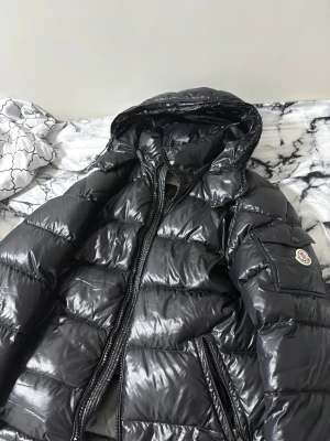Svart Moncler pufferjacka med huva - Säljer en svart pufferjacka från Moncler med glansig finish och huva. Jackan har quiltad design, dragkedja framtill och en ficka med Moncler-logga på ärmen. Perfekt för kalla dagar och har en riktigt snygg streetstil.
