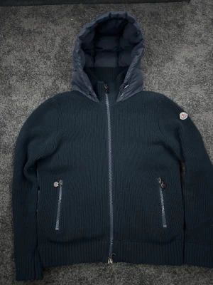 Marinblå Moncler wool cardigan med huva - Säljer en marinblå wool cardigan från Moncler med stickade ärmar och quiltad kropp. Jackan har en stor vadderad huva, två dragkedjeförsedda fickor och Moncler-logga på vänster ärm. Materialmix av stickad ull och syntet, perfekt för kyliga dagar.