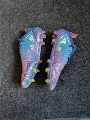 Adidas Messi blå fotbollsskor - Säljer ett par Adidas Messi fotbollsskor i blått med neonfärgade detaljer och texten 'Messi' i rosa över hela skon. Skorna har snörning, platt sula med dobbar i grönt och orange, samt Adidas-logga i gult. Ovandelen är i syntetmaterial och designen är riktigt sportig och modern.