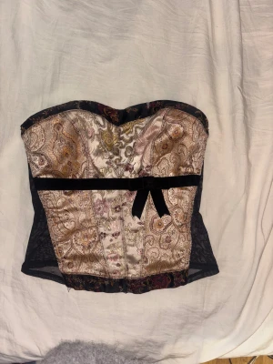 Paisley mönstrad korsett med rosett - Säljer en snygg korsett med paisley mönster i beige, guld och svart. Toppen har en svart mesh på sidorna och en söt sammetsrosett framtill. Stängs med hyskor i ryggen och har en croppad passform. Perfekt för dig som vill sticka ut!