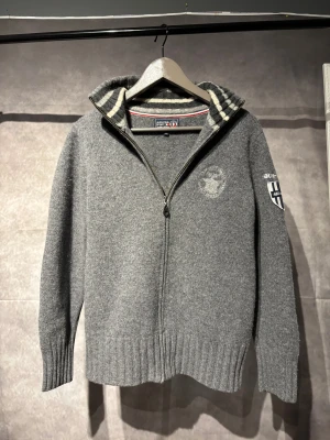 Jean Paul kofta - Snygg grå kofta från Jean Paul Sport i 100% ull med dragkedja framtill. Kragen har coola grå och vita ränder och broderade detaljer på bröstet och ärmen. Ribbstickade muddar och en normal passform gör den både chill och stilren.