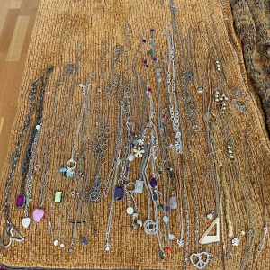 Stort halsbandspaket med hängen - Säljer ett stort paket med ett femtiotal halsband och armband i både silver- och guldfärgade kedjor. Flera halsband har coola hängen som hjärtan, blommor, stenar, geometriska former och färgglada detaljer i lila, blått, rött och grönt. Perfekt för dig som gillar att mixa och matcha olika stilar och uttryck.