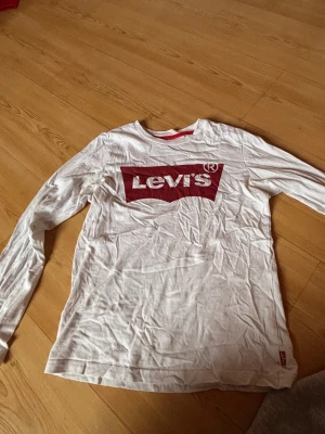 Vit långärmad Levi's tröja med röd logga - Vit långärmad tröja från Levi's med den klassiska röda loggan tryckt på bröstet. Tröjan har rund hals och är gjord i mjuk bomull, perfekt för chill dagar. Röd detalj med Levi's-märke i sidan. Enkel och clean stil som funkar året runt.