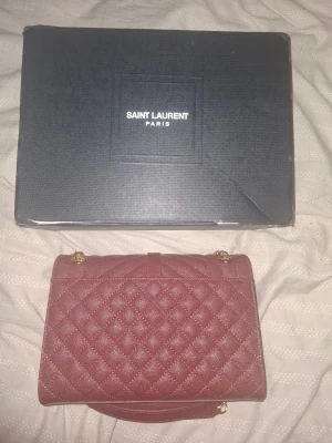 Quiltad röd Saint Laurent handväska - Säljer en snygg röd quiltad handväska från Saint Laurent med guldfärgade kedjor och YSL-logga framtill. Väskan är tillverkad i skinn och har en elegant, klassisk form. Perfekt för dig som vill ha en lyxig accessoar med ikonisk design.