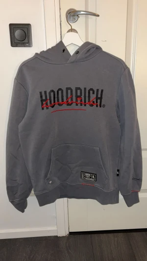 Grå Hoodie från Hoodrich med tryck - Hej! Snygg grå hoodie från Hoodrich i storlek S med svart och rött tryck på både fram- och baksidan. Tröjan har huva med snörning, stor magficka och coola detaljer som etikett och kontrastsömmar. Dock har den en liten vit fläck. Men den är Perfekt för dig som gillar streetwear och vill sticka ut.